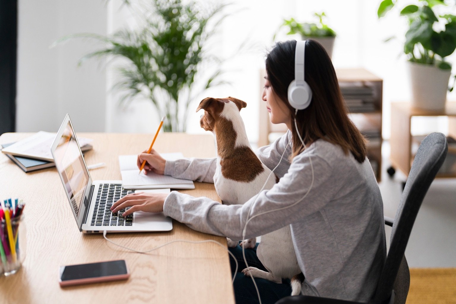A learner taking notes at a laptop in a home office, headphones on, with a small dog on their lap