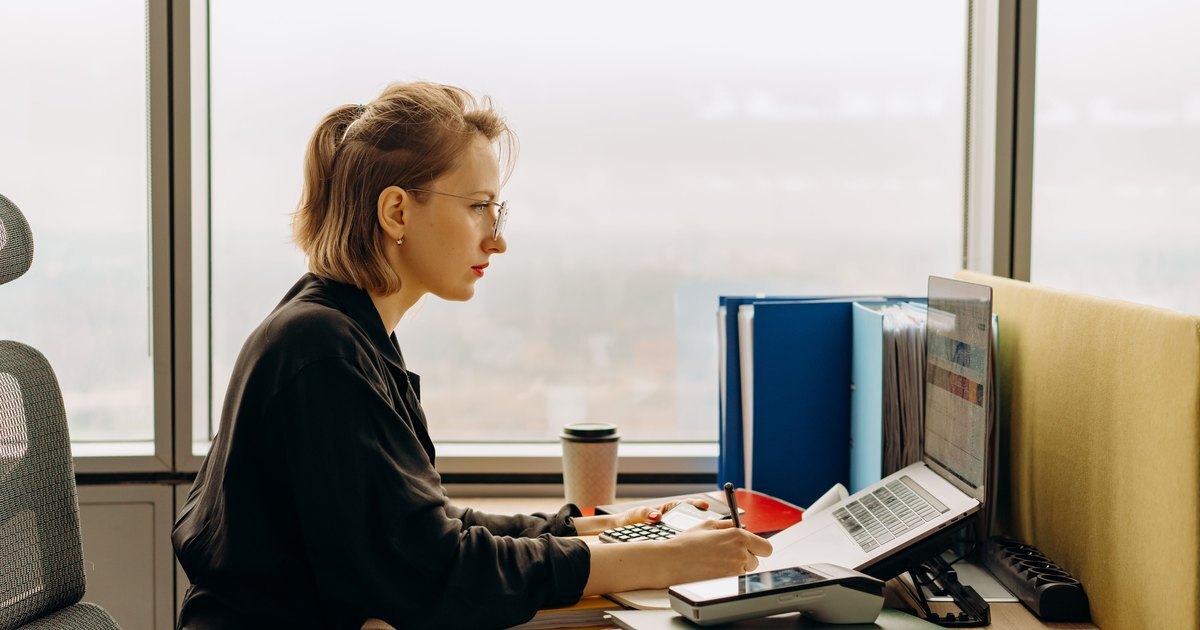 A professional at a desk by a window, working through figures with a calculator and laptop