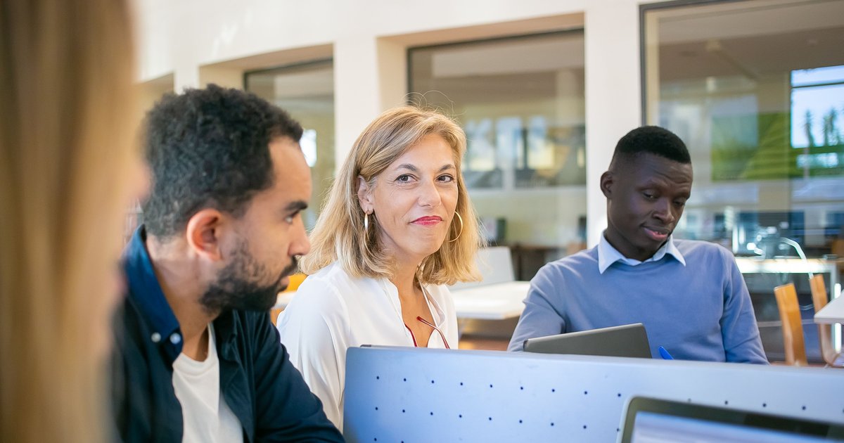 Three colleagues at a shared desk in a bright office, one smiling towards the camera