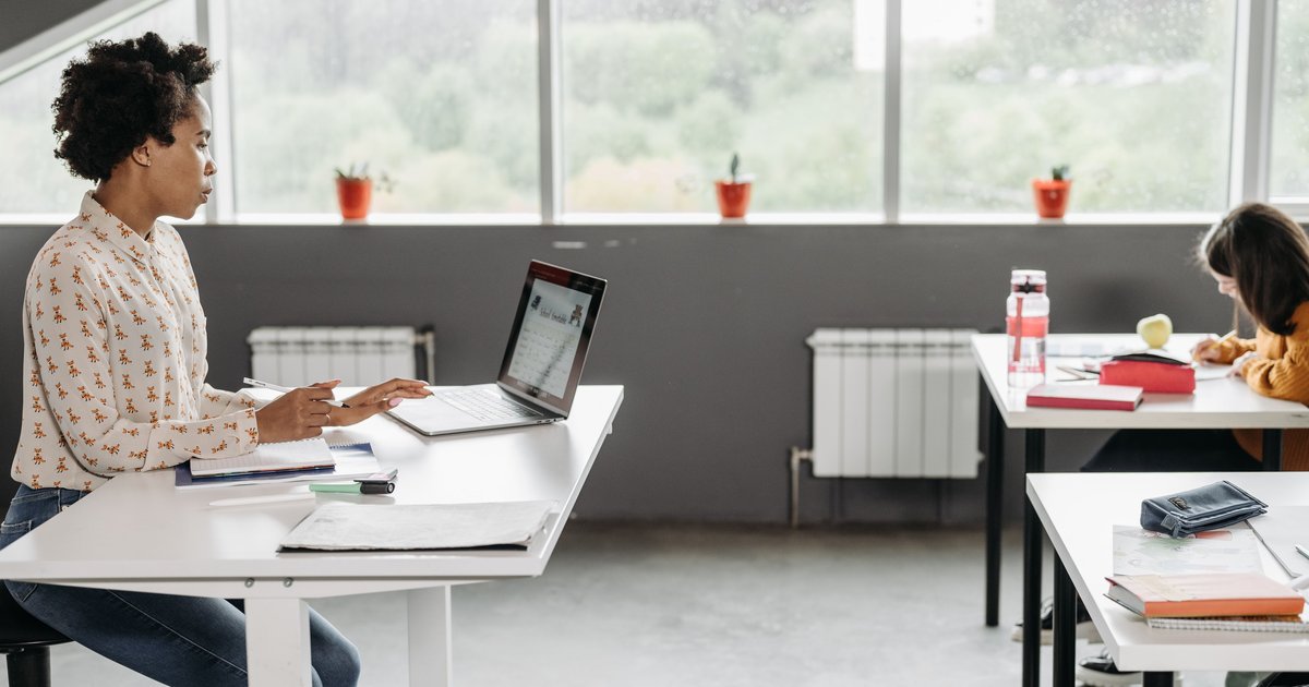 A teacher working on a laptop at the front of a classroom while a student writes at a desk