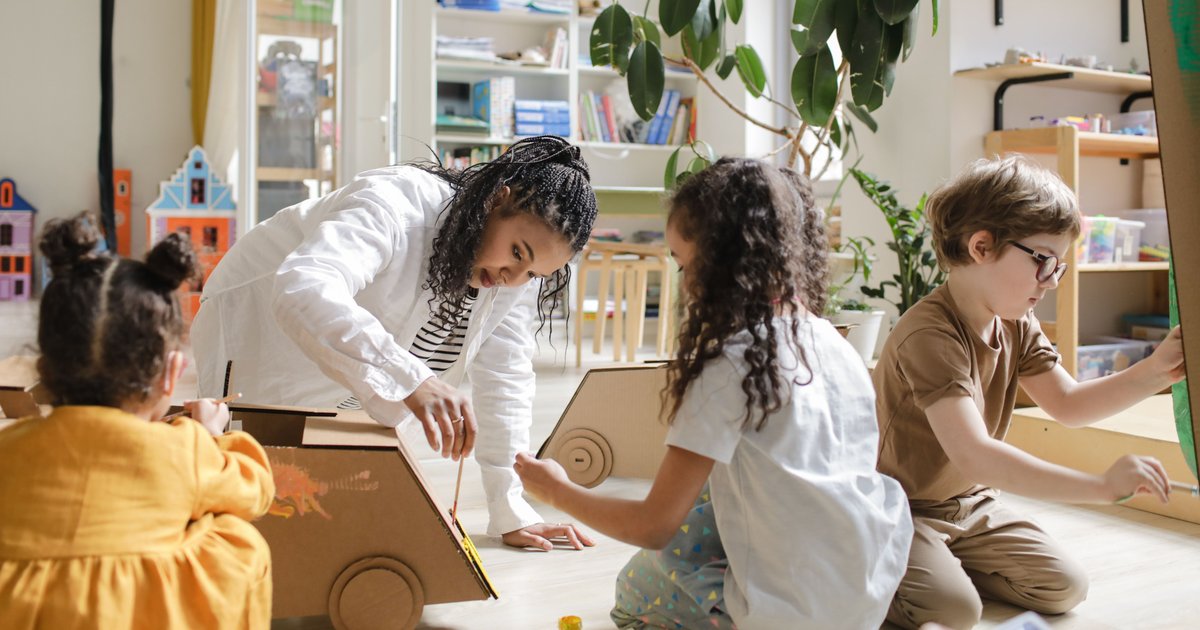 An educator crouches to help young children paint cardboard cars on a playroom floor