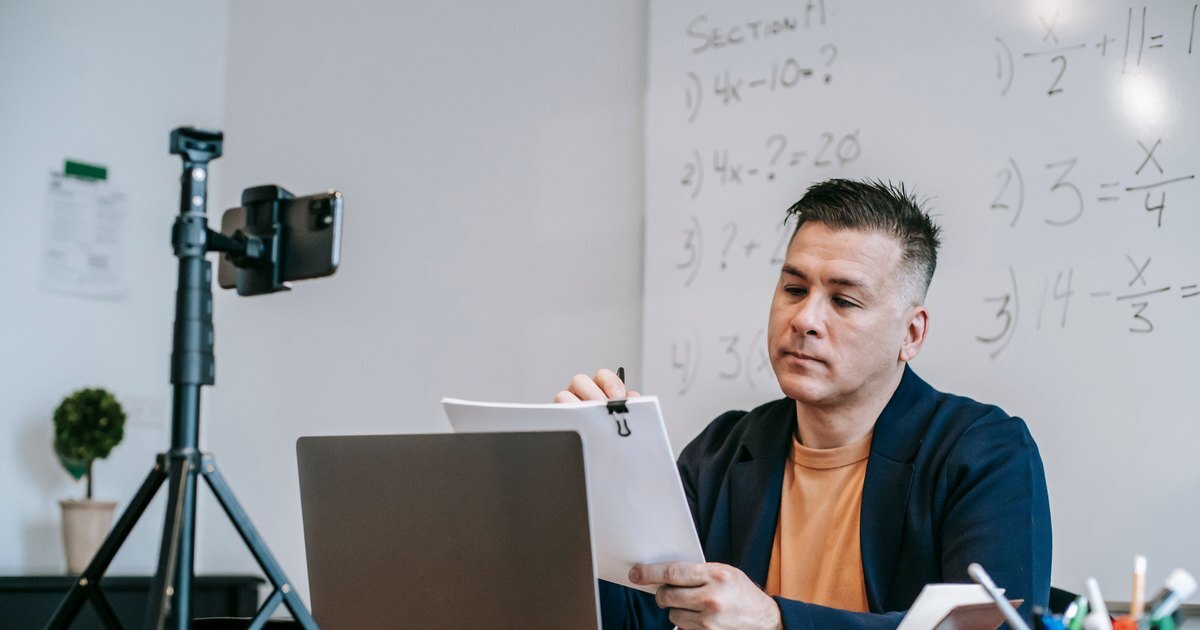 A lecturer at a desk with a laptop and notes, a phone on a tripod set up to record, a whiteboard behind him